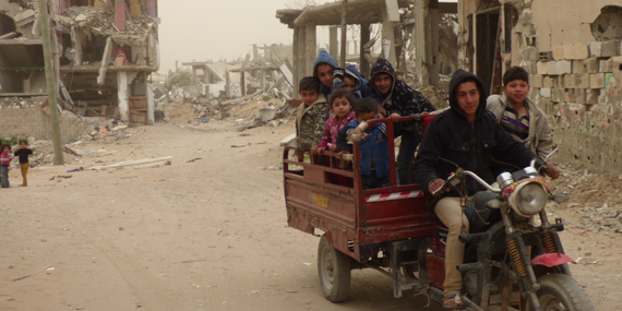 Children riding on a vehicle with damaged buildings from hostilities behind. Al Sha’af in At Tuffah area, Gaza. February 2015. Photo by OCHA