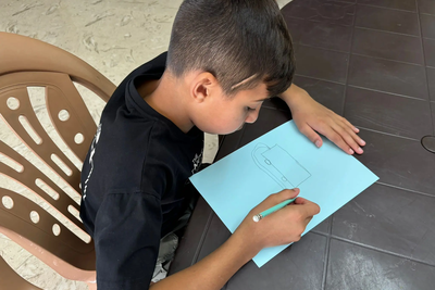 Kareem drawing a fire truck, sharing his dream of becoming a fireman. Three months ago, limited by his injury, he wished to be a carpenter. With nearly 90% recovery, his hopes have shifted back to firefighting, reflecting his progress and renewed sense of hope. Photo by © EJYMCA/Maha Sabbah