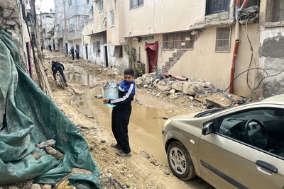 فتى فلسطيني يحمل الماء في شارع لحقت به أضرار جسيمة في مخيم طولكرم للاجئين عقب عملية إسرائيلية شهدت غارات شنّتها طائرات مسيرة عليه وتجريف الطرق فيه. تصوير مكتب الأمم المتحدة لتنسيق الشؤون الإنسانية