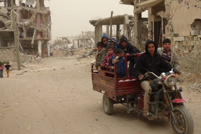 Children riding on a vehicle with damaged buildings from hostilities behind. Al Sha’af in At Tuffah area, Gaza. February 2015. Photo by OCHA